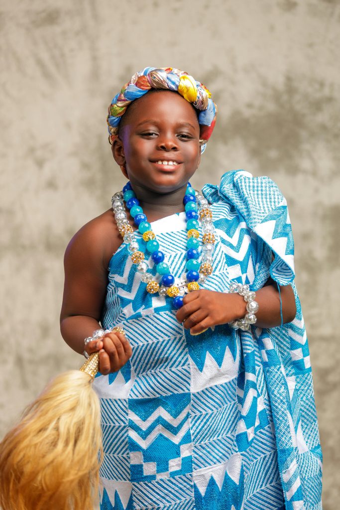 A happy child in vibrant tribal attire holding a broom, wearing a beaded necklace and headdress.